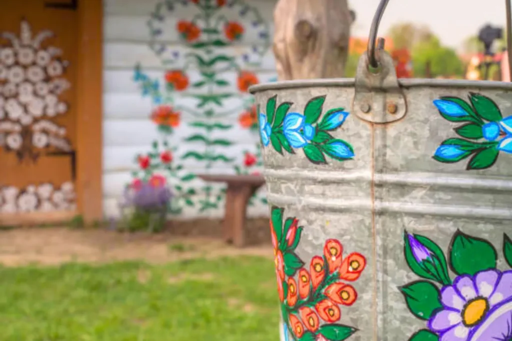 A vibrant painted metal bucket hangs prominently at the focal point, while in the background, an aged, painted house adds rustic charm to the scene.